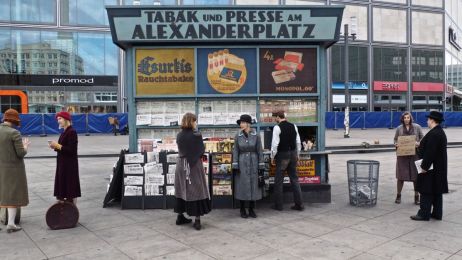 Black-and-white photo of a kiosk titled &raquo;Tobacco and Press&laquo; at Alexanderplatz, with three people, newspapers, cigarettes, and a bus in the background.